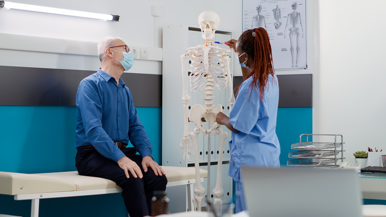 Medical assistant showing human skeleton bones to old patient at consultation during covid 19 pandemic. Nurse and senior man analyzing anatomy spinal cord to find orthopedic diagnosis 